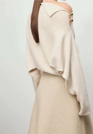 Femme aux longs cheveux raides portant un pull beige à épaules dénudées avec des boutons décoratifs et une jupe assortie, devant un fond uni.