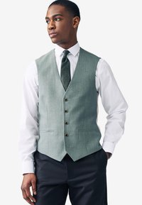 Gilet gris texturé à cinq boutons, porté par-dessus une chemise blanche et un pantalon foncé. Le gilet présente un col à encoche et des poches latérales.