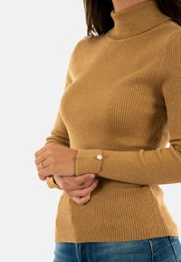 Maglione a collo alto a coste color cammello, con una silhouette aderente, una texture morbida e un accento di bottone argentato sul polsino.