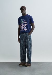 Μπλε μπλουζάκι με γραφικό σχέδιο που απεικονίζει έναν ροζ και πράσινο καρτούν χαρακτήρα, σε συνδυασμό με φαρδιά τζιν παντελόνια με υφή και μοτίβο.