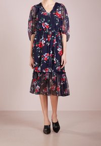 Mujer vistiendo un vestido midi floral de color azul marino con mangas abullonadas transparentes, cintura ceñida con un cinturón, dobladillo con volantes y mule negras de punta.