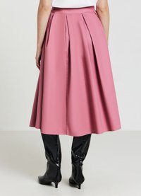 Una gonna rosa, lunghezza ginocchio, con pieghe, realizzata in un tessuto liscio. Indossata con stivali al ginocchio neri e lucidi. La vista da dietro mostra una cintura aderente.