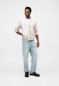 Veste beige clair zippée avec un motif texturé, associée à un jean bleu clair à jambes larges et des chaussures noires. Accessoires minimalistes.