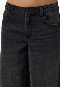Zwarte denim jeans met een gladde textuur, voorzien van een knoopsluiting, riemlussen en traditionele zakken. De stof vertoont subtiele vervaging.