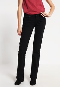 Schwarze, gerade Jeans aus Denim mit fünf Taschen und einem vorderen Knopfverschluss. Kombiniert mit einem figurbetonten roten Oberteil und schwarzen Stiefeln.