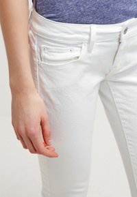 Jeans de mezclilla blanca con un diseño ajustado, que cuentan con dos bolsillos frontales y un cierre de botón metálico. El material parece suave con una ligera textura.