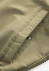 Gros plan d’un vêtement en tissu kaki avec une poche verticale et un bouton-pression argenté partiellement visible.