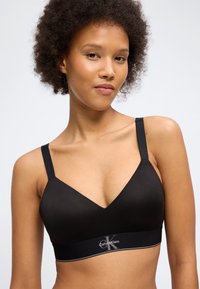 Νεαρή γυναίκα με σγουρά μαλλιά φορώντας ένα μαύρο σουτιέν Calvin Klein με ρυθμιζόμενους ιμάντες, πάνω σε ανοιχτόχρωμο φόντο.