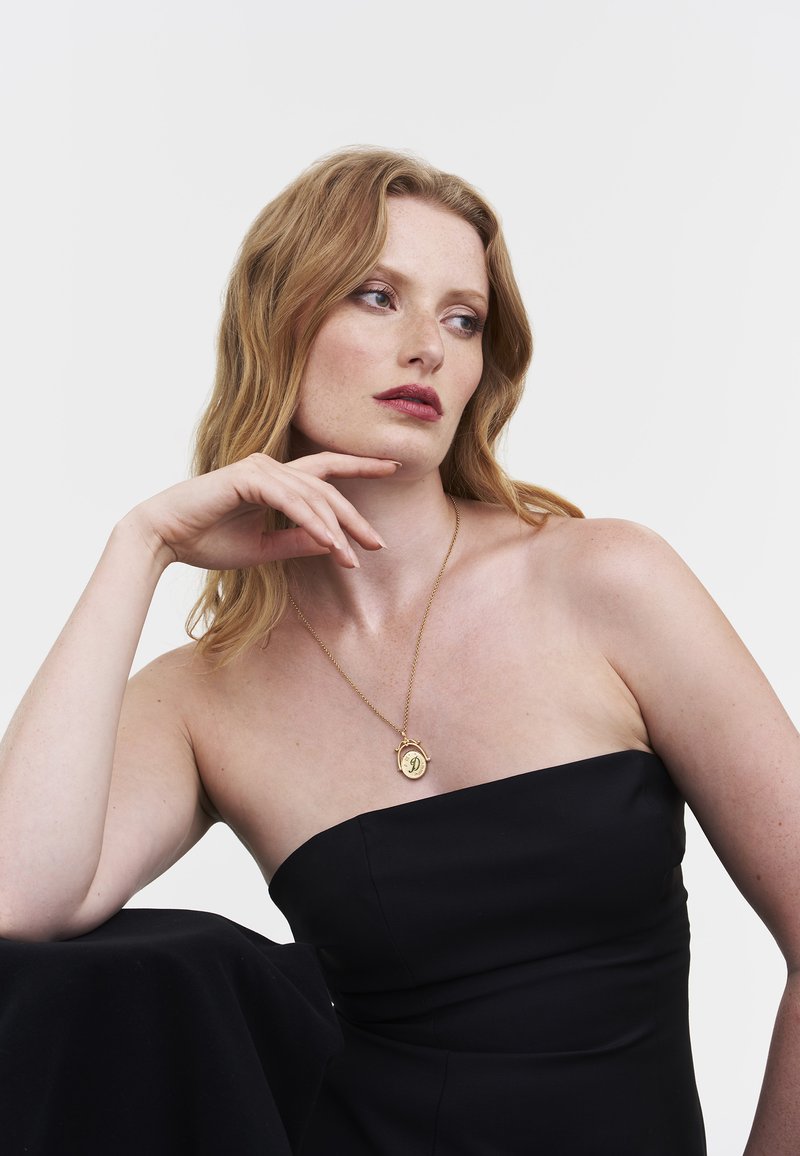 Kultainen riipuskaulakoru, jossa on pyöreä, kaiverretulla kirjaimella varustettu riipus, roikkuu sirosta ketjusta. Kaulakoru on pukeutuneena olkaimettoman mustan mekon päälle.