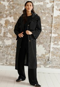 Svart vattentålig trench coat med huva, oversized passform, bälte och tryckknappsstängning. Bärs över svarta byxor och skor.