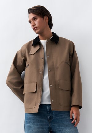 Junger Mann trägt eine beige Jacke mit schwarzem Kragen, weißem Hemd und blauen Jeans, steht vor einem schlichten weißen Hintergrund.