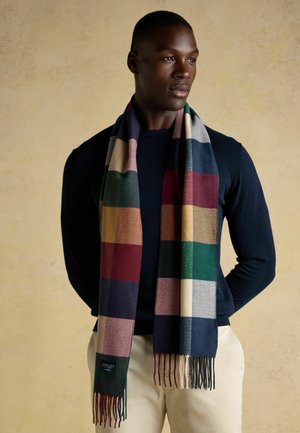 Man wearing dark navy sweater and beige pants with multicolored checkered scarf draped around his neck against a plain light background.