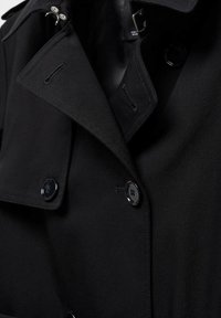 Schwarzer Trenchcoat mit doppelreihigem Design, glänzenden Knöpfen und strukturiertem Kragen. Das Gewebe ist glatt mit matter Oberfläche.