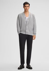 Grauer, knöpfbarer Cardigan mit lässiger Passform und gerippten Bündchen, kombiniert mit maßgeschneiderten schwarzen Hosen und schwarzen Schnürschuhen.