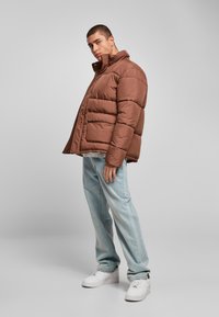 Brun oppustet jakke med høj krave, med syede sektioner og forlommer, parret med lyseblå jeans og hvide sneakers.