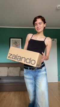 Scatola di cartone con la scritta "zalando" stampata in nero. Modello in un top nero senza maniche e jeans azzurri chiari, in piedi all'interno con uno sfondo di parete verde.