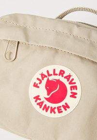 Sezione di una borsa Kånken beige di Fjällräven che mostra il patch del logo della volpe rossa e il tirante della cerniera con il nome del marchio su uno sfondo chiaro.