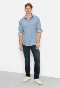 Mann steht an einer weißen Wand, trägt ein hochgekrempeltes hellblaues Hemd, dunkelblaue Jeans und weiße Sneakers mit beigen Sohlen.