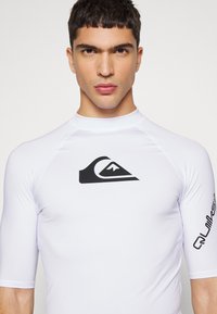 White rash guard with short sleeves, featuring a prominent black logo on the chest and black text along the right sleeve. Smooth, stretchy fabric.