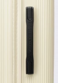 Detail van een witte harde koffer met een zwarte gestructureerde handgreep, een soepele rits en een verticale ribbelpatroon voor grip en duurzaamheid.
