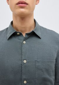 Άνδρας που φοράει σκούρο γκρι πουκάμισο με κουμπιά, γιακά και τσέπη στο στήθος, εμφανιζόμενος από τον λαιμό μέχρι το στήθος μπροστά σε απλό φόντο.