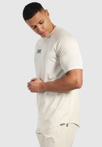 Camisa beige de manga corta con un patrón texturizado, con un logo negro en el frente y un dobladillo curvado. Llevada con pantalones de color claro.