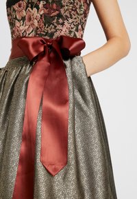 Bloemrijke bovenkant met een metallic gouden rok met een delicaat patroon. Brede bordeauxsatijnen lint geknoopt in een strik bij de taille.