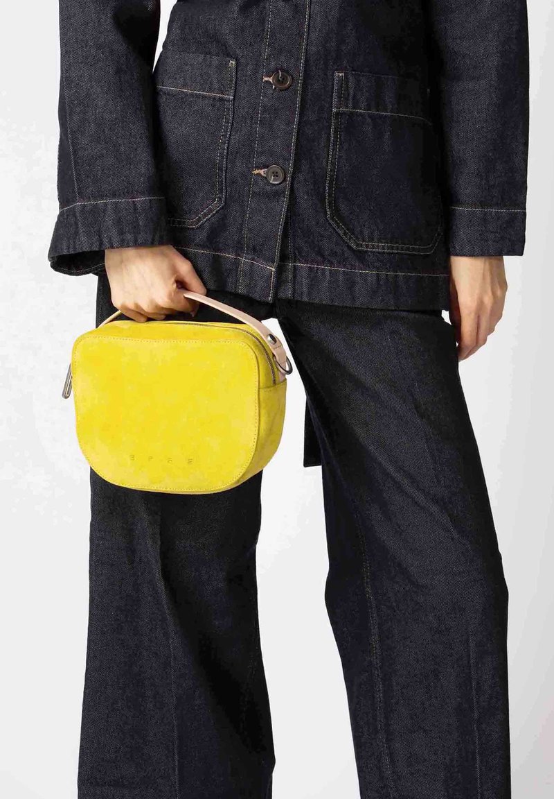 Geel suède handtas met een afgeronde vorm, leren handvat en geperst logo. Draagt een donker denim jack en broek.