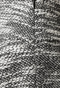 Zwart-witte geweven stof met een gestructureerd patroon. Kenmerkende steken en vezels zichtbaar, wat een gelaagd uiterlijk creëert.
