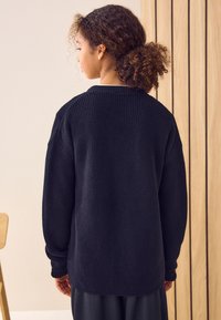 Črni pleteni pulover z ohlapno prileganostjo, rebrastimi manšetami in teksturiranim vzorcem. Hrbtni del je preprost, brez vidnih logotipov ali okraskov.