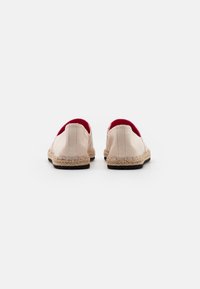 Bottes slip-on en toile beige avec intérieur rouge, dotées d'une semelle en jute tissé et d'une base en caoutchouc pour l'adhérence. Design simple avec une texture douce.