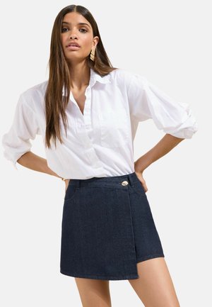Vrouw met lang steil haar draagt een wit blouse met knopen en opgerolde mouwen en een donkerblauwe denim wikkelrok met gouden knoop.