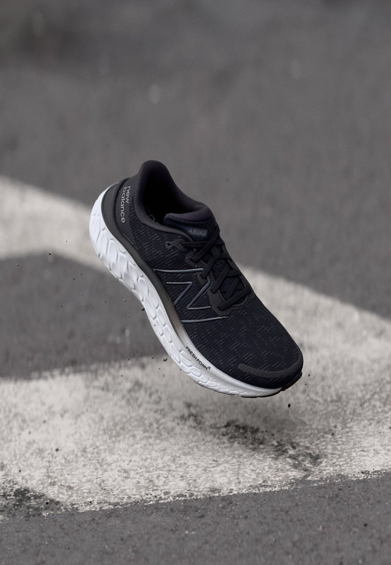 Scarpa sportiva nera con tomaia texturizzata, intersuola bianca e dettagli grigi, dotata di lacci e dettagli del logo, mostrata in volo sopra una strada.