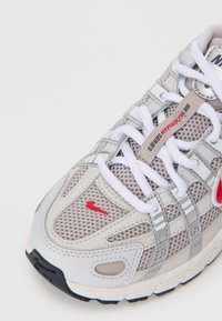 Sive mrežaste tenisice s bijelim vezicama i crvenim Nike logom. Imaju srebrne detalje i zaobljeni vrh, dizajnirane za udobnost i potporu.