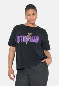 Černé oversized tričko s leopardím potiskem blesku a výrazným fialovým nápisem "STUDIO." Měkká bavlněná tkanina s krátkými rukávy.