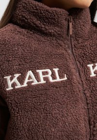 Brun fleecejakke med høj krave, der har cremefarvet "KARL" broderi, lynlås lukning og en tekstureret, blød stofflade.