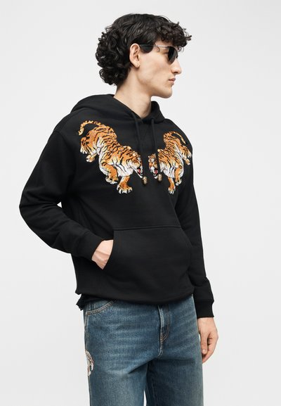 Νεαρός άνδρας που φοράει μαύρο φούτερ με κουκούλα με δύο κεντημένες τίγρεις στο στήθος, μπλε τζιν με μπαλώματα τίγρης στον μηρό και σκούρα γυαλιά ηλίου, κοιτάζοντας προς τα δεξιά.