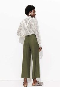 Haut en dentelle avec des détails froncés, associé à un pantalon large vert olive. Le modèle porte des sandales à imprimé léopard. Vue de dos, sur un fond uni.