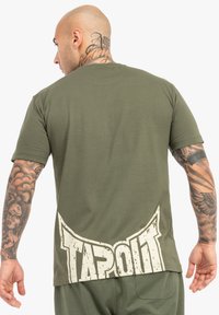 OLIVOVO ZELENÁ KÚDOLENÁ TIŠTOVKA S KRÁTKYMI RUKÁVMI, S VEĽKÝM BIELYM LOGOM "TAPOUT" NA ZADNEJ STRANE, HLASNEJŠIE TEXTÚRY A UVOĽNENÝM STRIHOM.