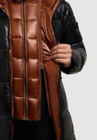 Pufjakke med en skinnende sort ydre og en kontrastfyldt glat brun inderside. Har kviltet tekstur og dobbelte lynlåse.