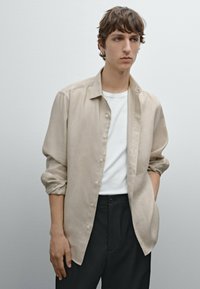 Beige Leinenhemd mit Knöpfen und lässigem Schnitt, getragen über einem weißen T-Shirt, kombiniert mit schwarzen Hosen; angenehme Textur, lässiges Design.