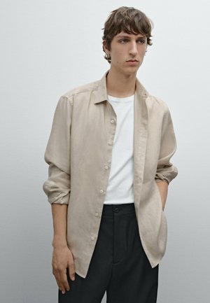 Beige linned skjorte med knapper og afslappet pasform, båret over en hvid T-shirt, sammen med sorte bukser; glat tekstur, afslappet design.