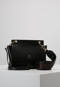 Czarna skórzana torebka Aigner z złotymi akcentami i odpinanym szerokim paskiem z wycięciami w kształcie łuku, wyeksponowana na białym piedestale.