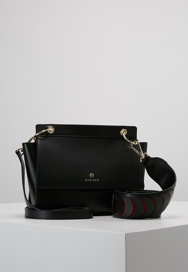 Czarna skórzana torebka Aigner z złotymi akcentami i odpinanym szerokim paskiem z wycięciami w kształcie łuku, wyeksponowana na białym piedestale.