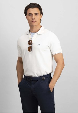 Mężczyzna w białej koszulce polo z krótkim rękawem i małym logo, granatowych spodniach, plecionym pasku i okularach przeciwsłonecznych zawieszonych na kołnierzu koszulki, z rękami w kieszeniach.