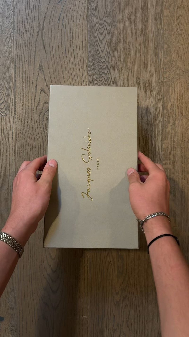 Светлосив квадрат с златисто скриптово надпис "Jacques Soufflé Paris." Гладка матова текстура, държан отгоре с две ръце, почиващ на дървена повърхност.