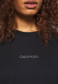 Sudadera de marina con cuello acanalado, que presenta un logo bordado en blanco "CALVIN KLEIN" en la parte delantera. Textura suave y diseño ajustado.