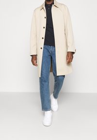 Man som går klädd i en beige knappad kappa, svart pikétröja, blå jeans och vita sneakers mot en enkel ljus bakgrund.