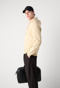 Felpa con cappuccio giallo chiaro, cappellino nero, pantaloni scuri, porta una borsa nera. Tessuto morbido, vestibilità rilassata, dettagli di cucitura sottili.