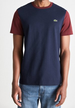 Mann in navyblauem T-Shirt mit burgunderfarbenen Ärmeln und Kragen, kombiniert mit blauen Jeans, steht vor einem einfarbigen Hintergrund.